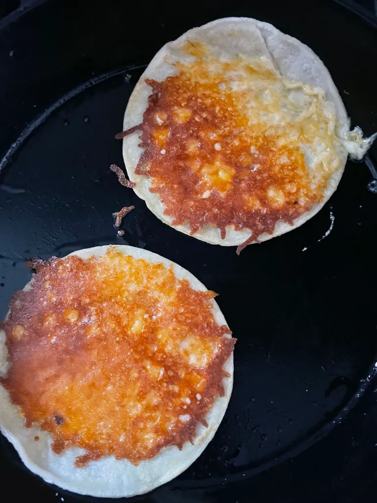 Two corn tortillas flipped cheese-side up in the cast iron skillet, showing the golden, crispy browned cheese crust