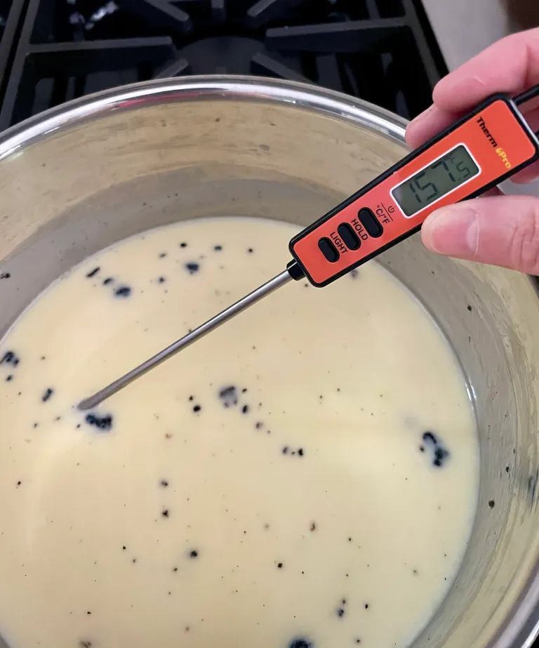 An instant-read thermometer reading 157°F being used to check the temperature of the vanilla cream mixture in a saucepan, with vanilla bean specks visible throughout