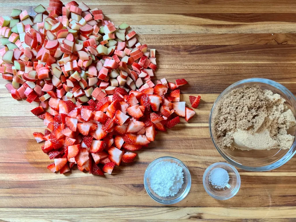 Flat lay of strawberry-rhubarb filling ingredients on a wood cutting board: chopped fresh rhubarb, quartered fresh strawberries, a bowl of brown sugar, cornstarch, and kosher salt