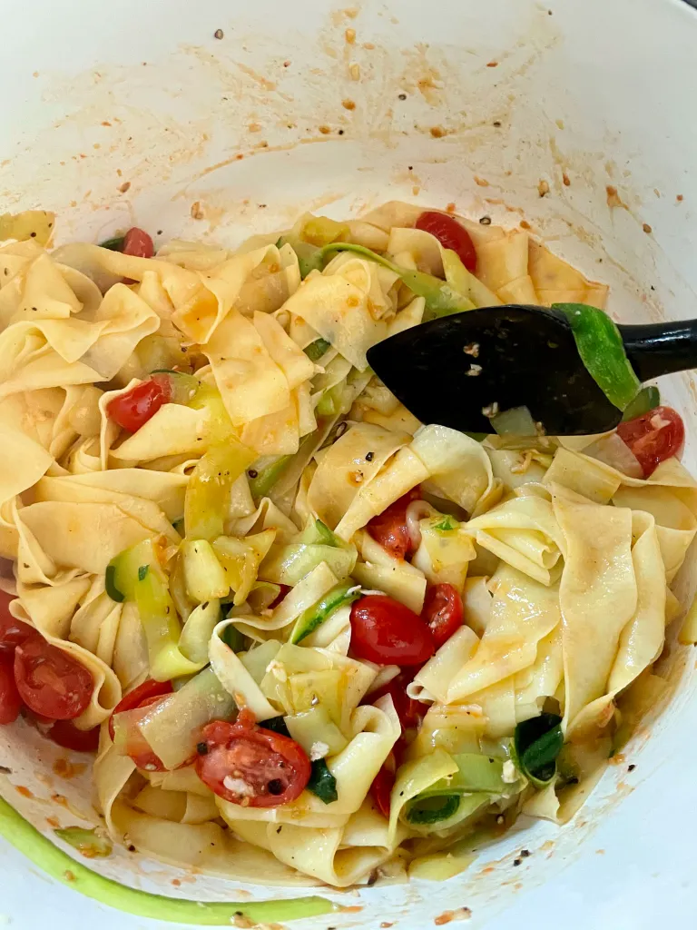 Pappardelle gently tossed together with zucchini ribbons, burst tomatoes, and garlic in the white Dutch oven using black tongs, coated in a light tomato-garlic sauce