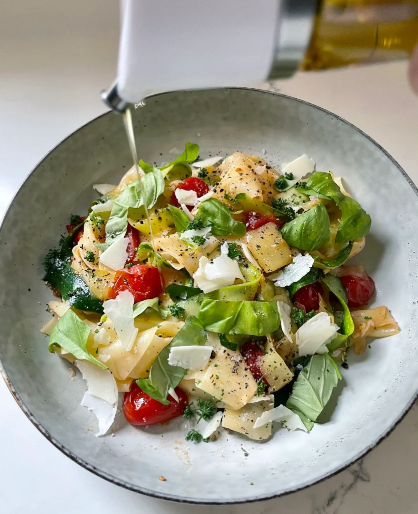 Finished summer pasta in a speckled gray bowl being drizzled with olive oil from a bottle, topped with shaved Parmesan, fresh basil, and parsley with a bottle of olive oil and fork visible