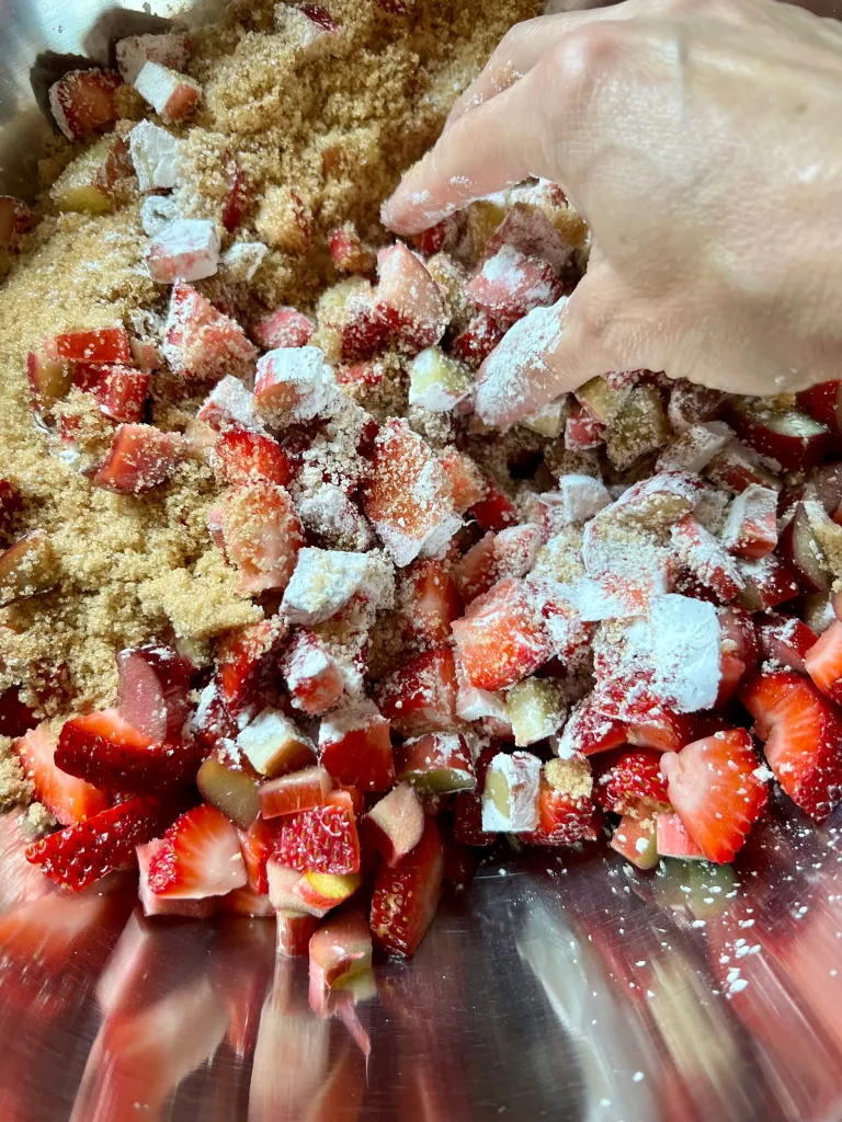 Chopped strawberries and rhubarb being tossed with brown sugar and cornstarch in a large stainless mixing bowl by hand