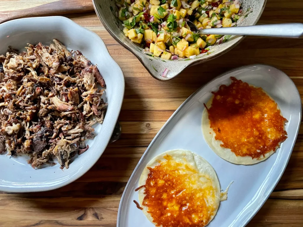 Overhead taco assembly station with a white dish of crispy shredded smoked pork shoulder, two cheesy corn tortillas on a white oval platter, and a bowl of chipotle pineapple salsa with a spoon