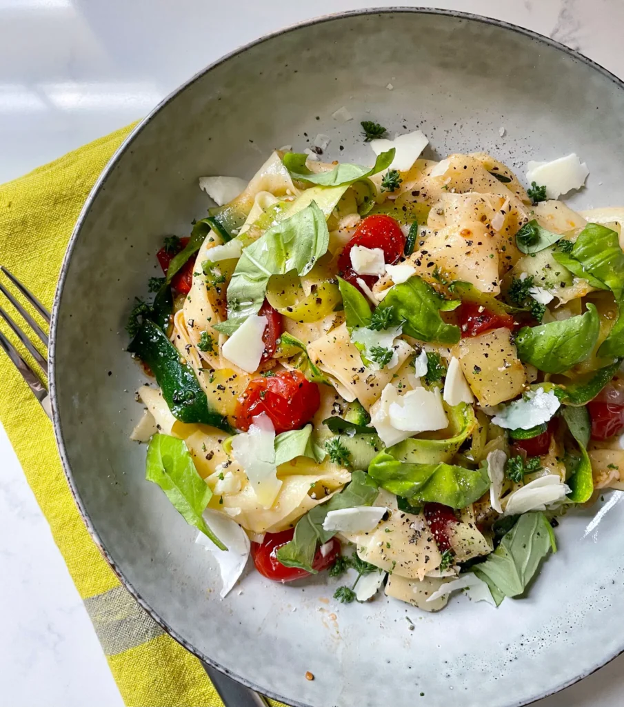 Overhead shot of the finished summer pasta with zucchini and tomatoes in a speckled gray shallow bowl on a yellow linen napkin, topped with shaved Parmesan, fresh basil, parsley, and cracked black pepper