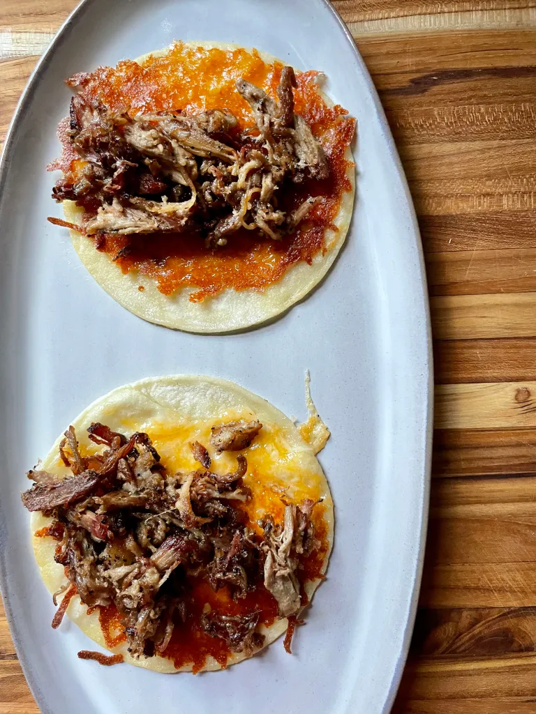 Two cheesy corn tortilla tacos topped with crispy shredded pork on a white oval platter on a wood cutting board, before salsa is added