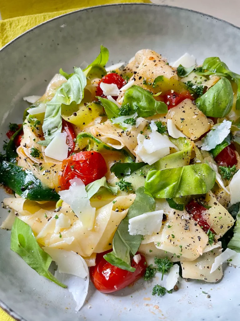 Close-up of summer pasta with zucchini and tomatoes in a speckled gray bowl on a yellow napkin, showing the silky pappardelle ribbons, burst tomatoes, zucchini strips, shaved Parmesan, and fresh basil and parsley garnish