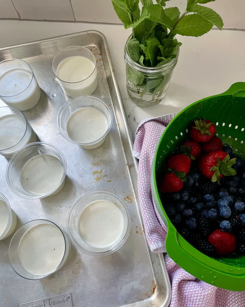 Set panna cottas in clear cups and glasses on a rimmed sheet tray alongside a colander of fresh strawberries, blueberries, and blackberries and a mason jar of fresh mint, ready for topping