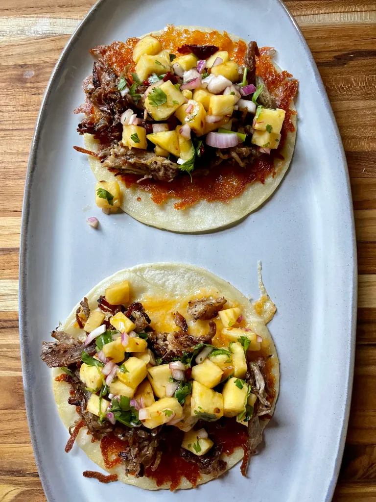 Two finished pork tacos on a white oval platter topped with chipotle pineapple salsa, showing diced pineapple, red onion, and cilantro over crispy pork and a golden cheese crust