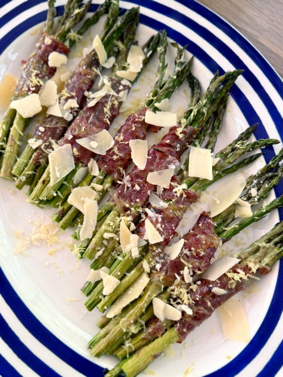 A close-up overhead view of roasted asparagus and prosciutto bundles on a white navy-striped oval platter, the crispy prosciutto caramelized and golden, topped with generous shavings of Parmesan and fresh lemon zest.