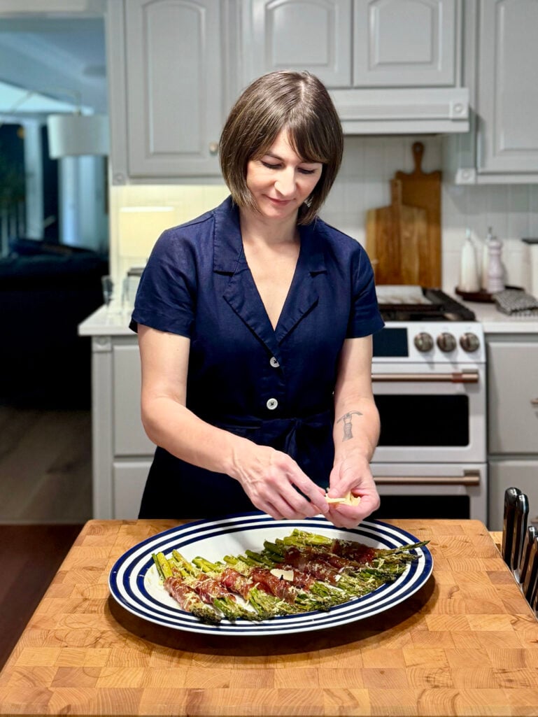 Kelly stands at a butcher block kitchen counter, smiling as she carefully adds shaved Parmesan to a platter of roasted asparagus and prosciutto bundles, wearing a navy linen shirtdress, with gray cabinets and a stainless range visible behind her.