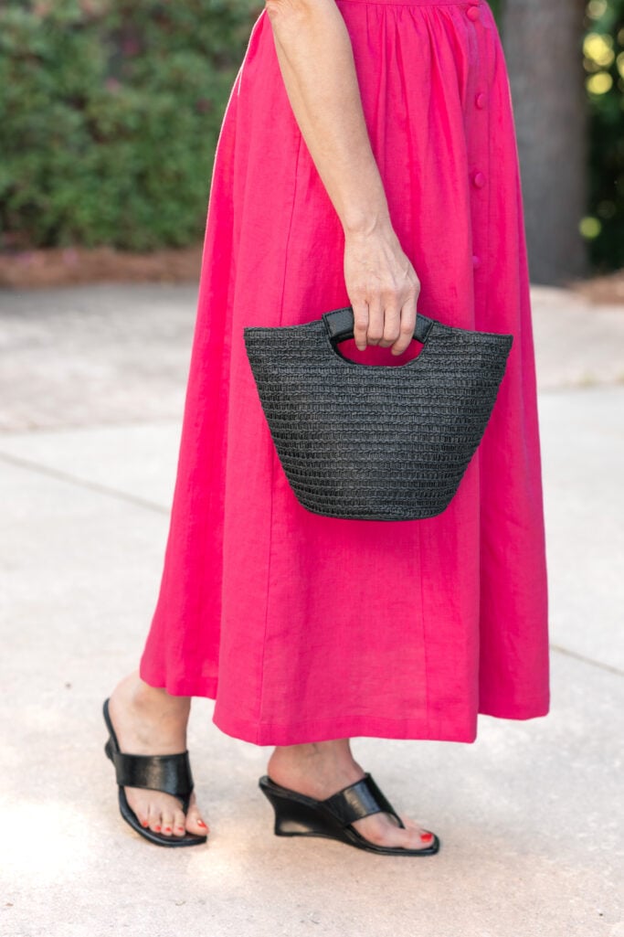 Close-up of black straw mini bucket bag and black thong wedge sandals styled with hot pink linen maxi skirt — petite summer accessories