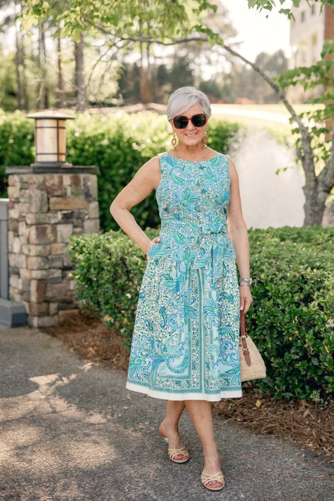 Woman over 50 with silver hair standing outdoors wearing a blue and green paisley fit and flare midi dress with a self-tie waist detail and white hem border, styled with large wood drop earrings, a natural woven shoulder bag with cognac leather handles, tortoise sunglasses, and nude woven kitten mule sandals — a polished Mother's Day dress look for women over 50 in regular sizing.