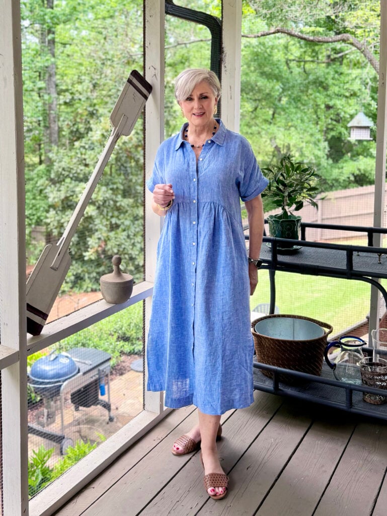 Beth stands on a screened porch in a periwinkle blue linen shirtdress with a smocked waist, woven leather slide sandals, a beaded necklace, and gold hoop earrings, one hand raised, with lush green trees visible through the porch screens behind her.