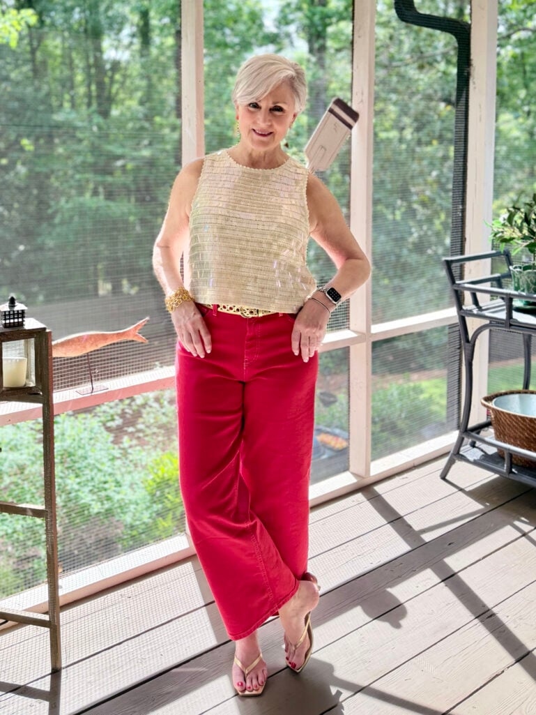 Beth stands on the screened porch in a cream iridescent sequin shell top, deep red wide-leg crop jeans with a gold braided leather belt, and gold thong kitten heel sandals, one hand on her hip, smiling at the camera with lush green trees visible through the porch screens behind her.