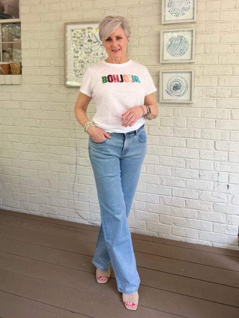 Beth stands on the screened porch against a white brick wall, one hand in her pocket, wearing a white sequin Bonjour graphic tee, light wash rhinestone-embellished wide-leg jeans, and nude heeled slide sandals, smiling at the camera with the Georgia map print visible behind her.
