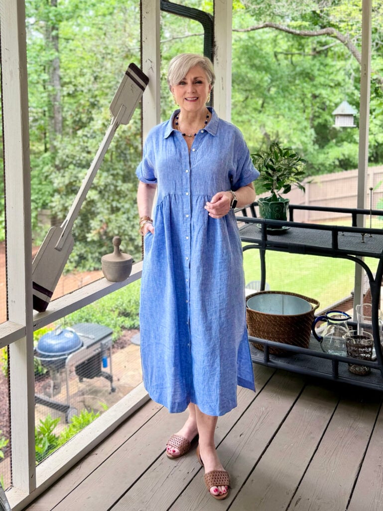 Full-length view of Beth on the screened porch in her periwinkle linen shirtdress and woven sandals, smiling with both hands clasped lightly in front, green trees and a bar cart visible through the porch screens in the background.