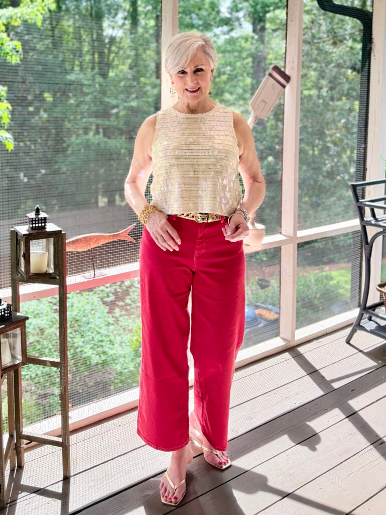 Full-length view of Beth on the screened porch in her sequin shell top, red wide-leg crop jeans, gold belt, and gold kitten heel sandals, both hands resting at her hips, with porch lanterns and greenery visible behind her.