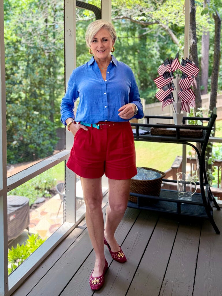 Full-length view of Beth in a blue linen shirt and red cuffed shorts, hand in pocket, smiling on the screened porch with American flags and a bar cart in the background.