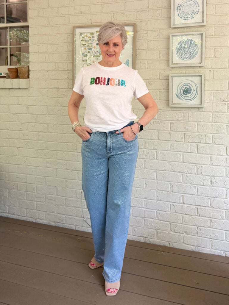 Full-length view of Beth on the porch in her white Bonjour sequin tee, rhinestone wide-leg denim, and nude slide sandals, both hands in her pockets, with blue abstract art prints and the Georgia map framed on the white brick wall behind her.