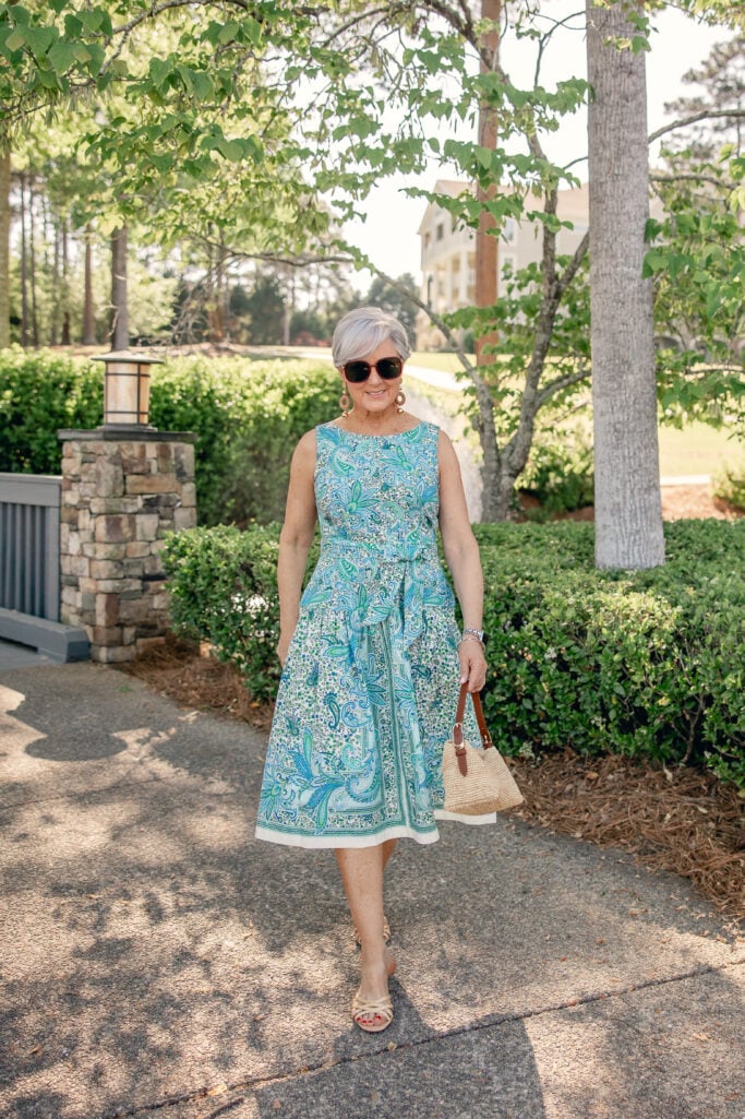 Woman over 50 with silver hair walking outdoors in a blue and green paisley fit and flare midi dress with a self-tie waist, wood drop earrings, tortoise sunglasses, a natural woven bag with cognac leather strap, and nude kitten mule sandals — full-length view of the Mother's Day dress in regular sizing on a woman over 50.
