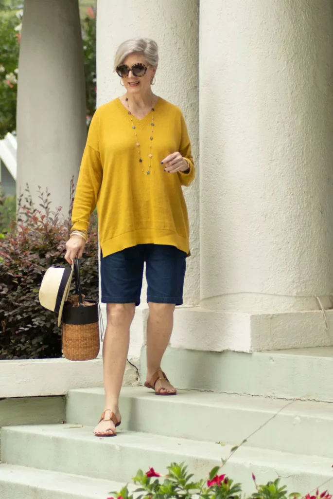 Woman over 50 with silver hair wearing a mustard yellow lightweight summer sweater with dark denim Bermuda shorts, tan T-strap sandals, and a straw bucket bag with tortoise sunglasses and a long beaded necklace — an easy tonal Bermuda shorts outfit idea for women over 50.