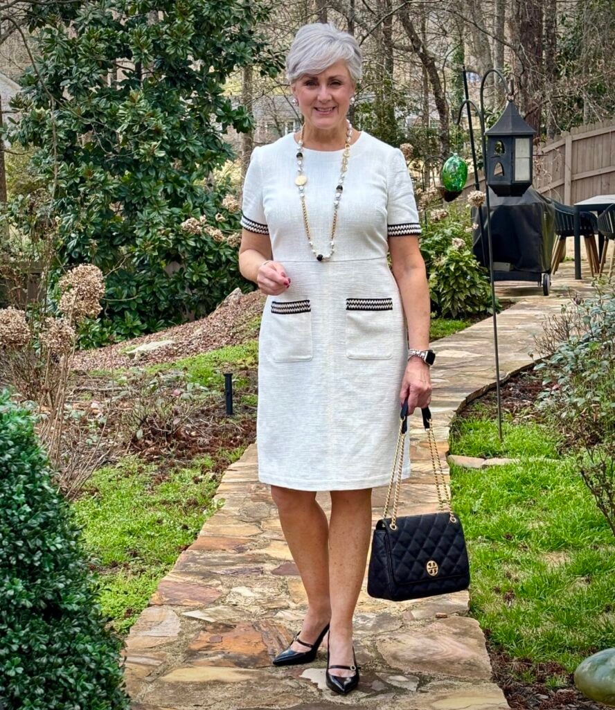 Woman over 50 with silver hair wearing an ivory cotton bouclé short-sleeve shift dress with black ribbon trim on the cuffs and front pockets, styled with a pearl and gold link statement necklace, pearl earrings, black Mary Jane pumps, and a black quilted chain-strap handbag — a polished spring or summer occasion dress outfit idea for women over 50.