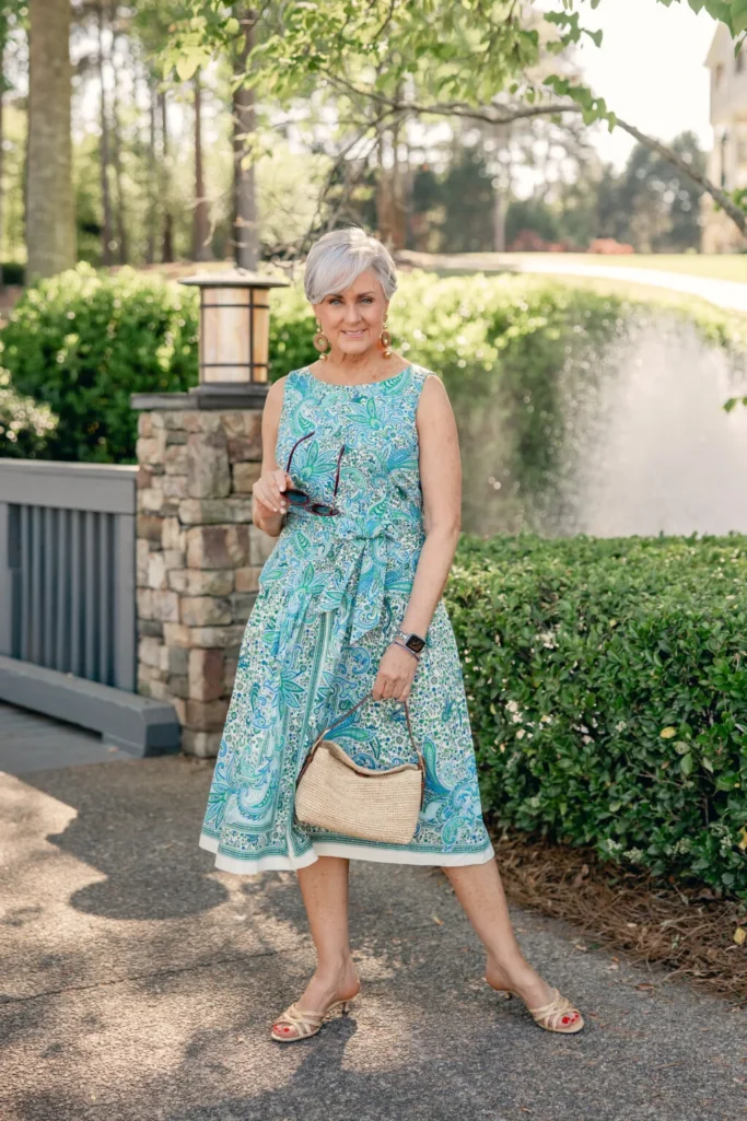 Woman over 50 with silver hair wearing a sleeveless blue and green paisley print cotton poplin fit-and-flare midi dress with a defined waist and full skirt, carrying a natural raffia handknotted shoulder bag with strappy gold sandals and wood drop earrings — a flattering summer cotton dress outfit idea for women over 50.