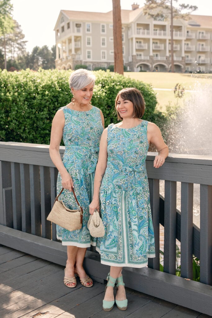 Two women laughing together on a wooden bridge, both wearing the same blue and green paisley fit and flare midi dress — the woman on the left (over 50, silver hair) in regular sizing with gold drop earrings and a woven shoulder bag, and the woman on the right in petite sizing with a cream woven clutch and mint wedge espadrilles — a candid Mother's Day dress comparison in petite and regular.