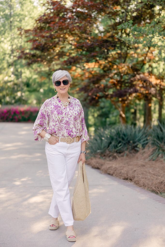 Woman over 50 wearing white jeans with a purple paisley blouse, raffia woven belt, beaded necklace, espadrille platform slides, and straw tote — summer weekend outfit with white jeans for women over 50.