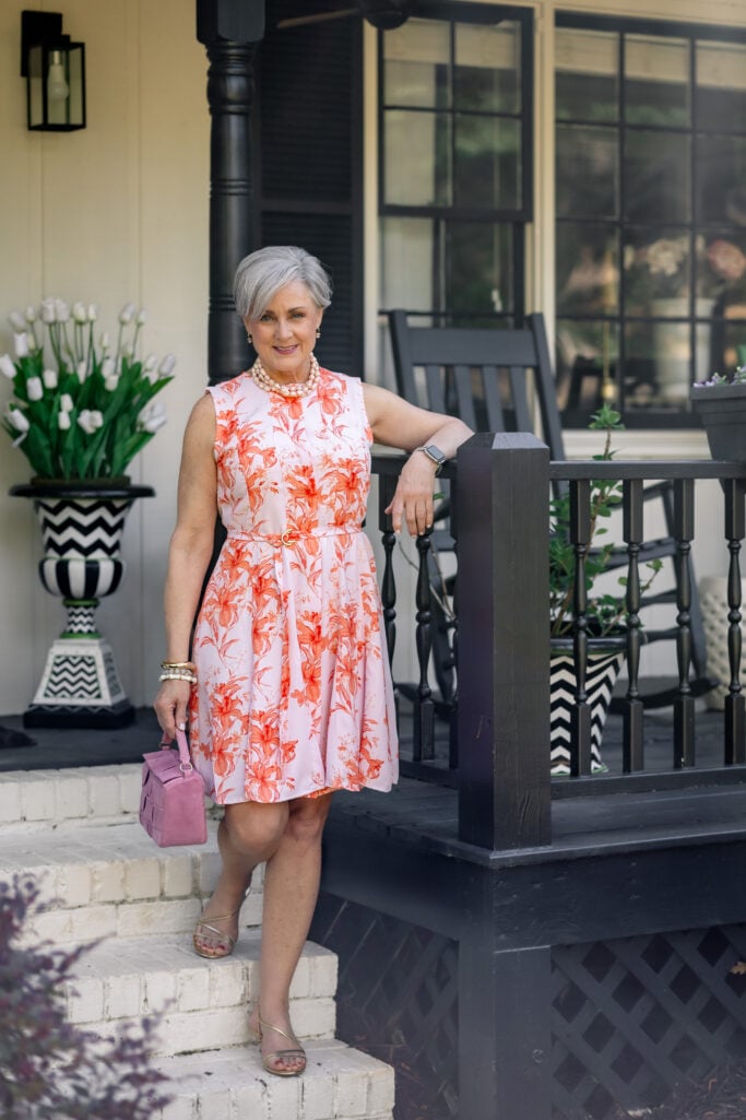 Peony pink and coral floral fit-and-flare midi dress with tie waist, pearl statement necklace, and pink suede top-handle bag.