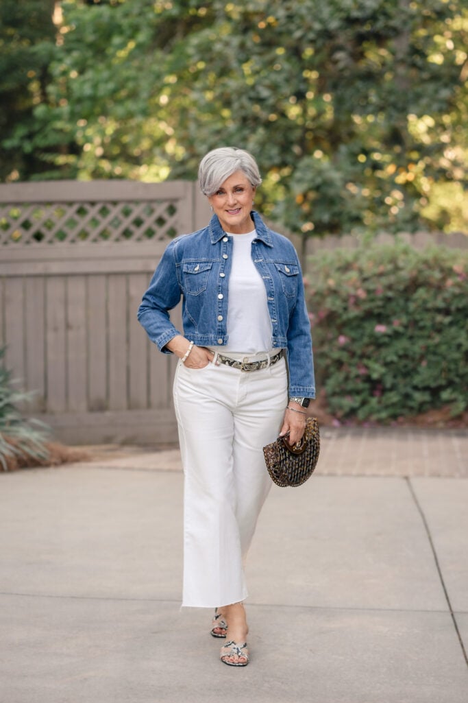Woman over 50 wearing white wide-leg cropped jeans with a cropped denim jacket, white tee, snake-print belt, and snake-print heeled sandals — casual lunch outfit with white jeans for women over 50.