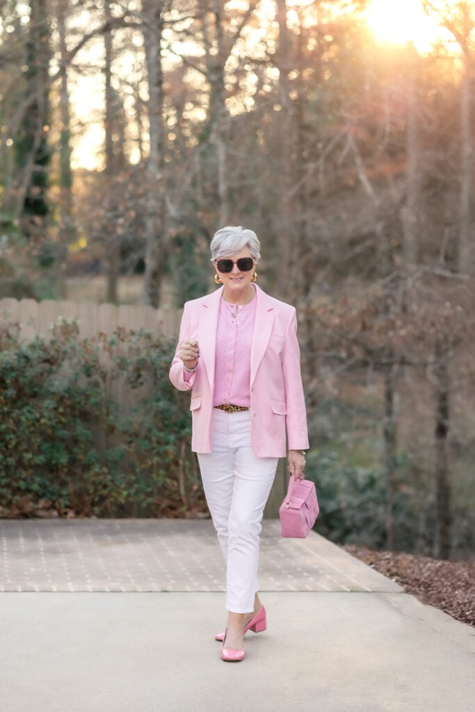 Woman over 50 wearing white jeans with a pink blazer, pink cardigan, leopard belt, pink patent slingback heels, and pink woven handbag — date night outfit with white jeans for women over 50.
