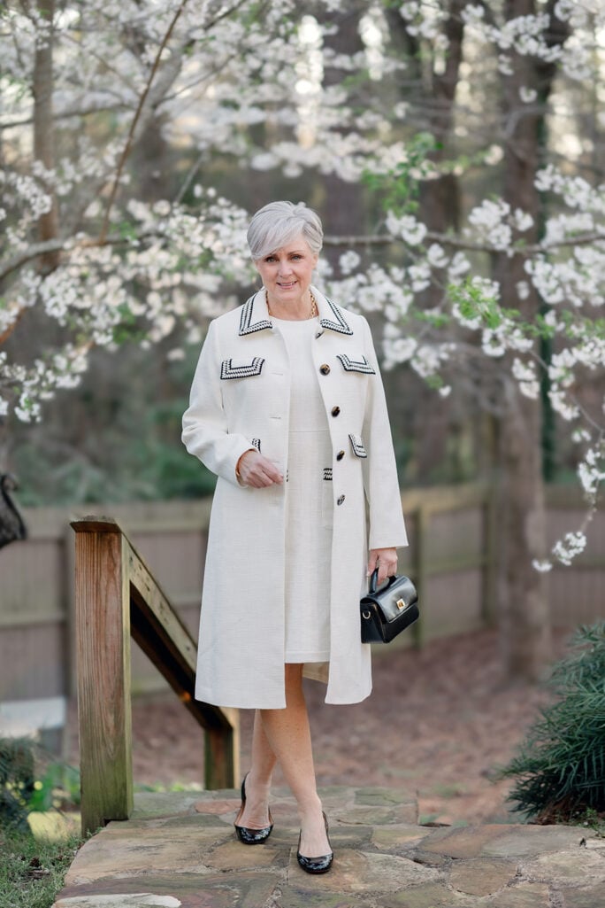 Woman over 50 with silver hair wearing a cream cotton bouclé short-sleeve shift dress with a matching cream duster coat, styled with black patent Louboutin pumps, a gold chain link necklace, gold cuff bracelet, and a small black structured top-handle bag — an elegant monochromatic cream outfit idea for spring or summer occasions for women over 50.