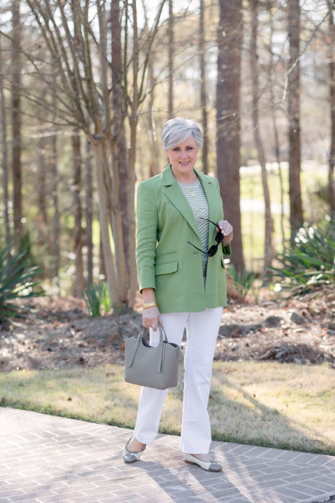 Woman over 50 wearing white flared jeans with a green blazer, green and white striped tee, gray leather tote, and two-tone ballet flats — spring work outfit with white jeans for women over 50.
