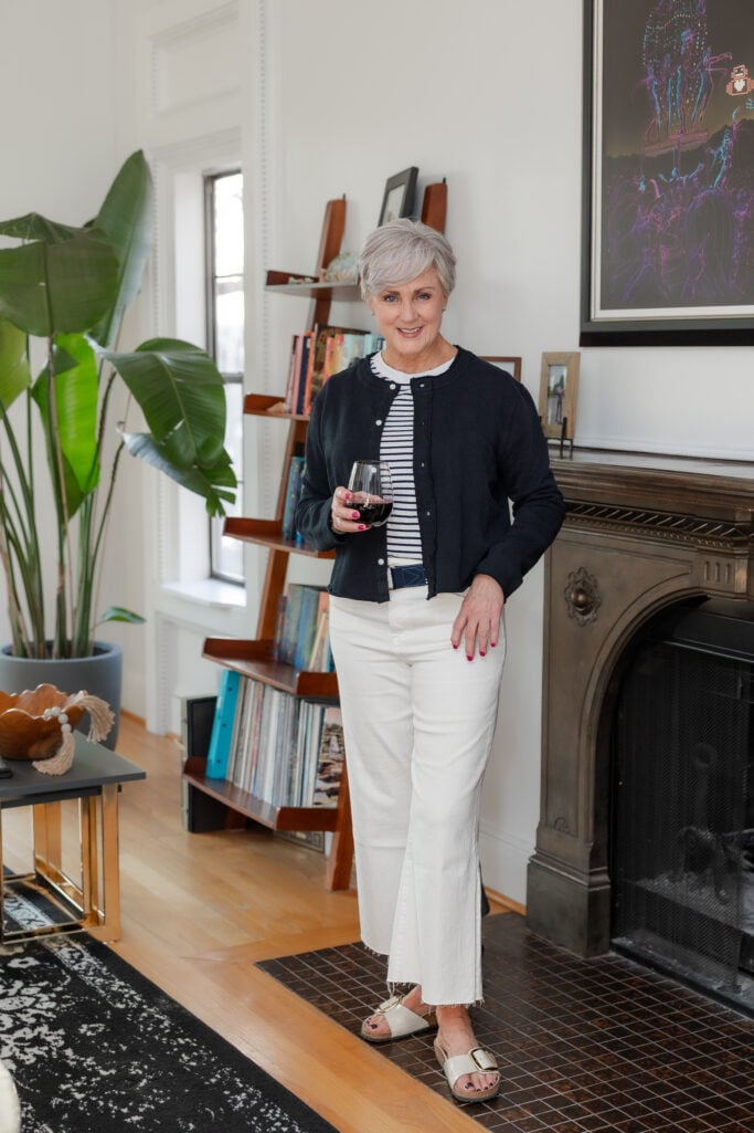 Woman over 50 wearing white cropped flared jeans with a navy cardigan, navy and white striped tee, and white leather slides, holding a glass of red wine — relaxed evening at home outfit with white jeans for women over 50.