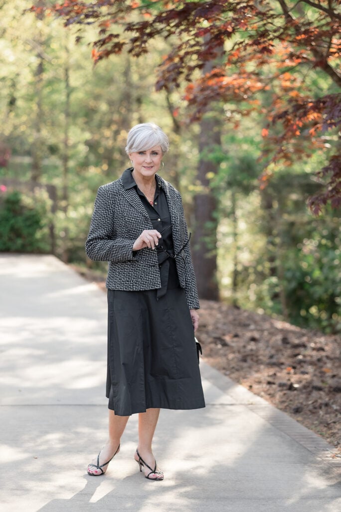 Woman over 50 with silver hair wearing a black cotton midi shirtdress layered under a black and white tweed jacket with strappy black sandals, gold hoop earrings, a delicate red heart pendant necklace, and stacked gold bangles — an elevated summer shirtdress outfit idea for women over 50.