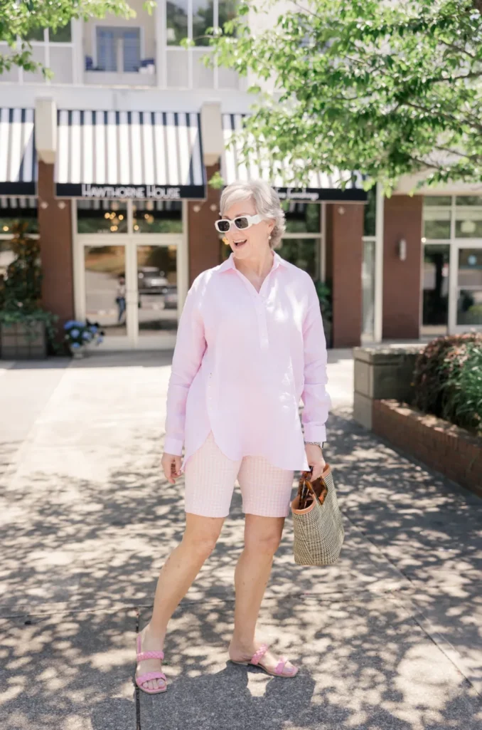 Woman over 50 with silver hair wearing pale pink gingham Bermuda shorts with a soft pink linen popover shirt, pink braided slide sandals, and a woven top-handle bag with white sunglasses — a soft neutral Bermuda shorts outfit idea for women over 50.