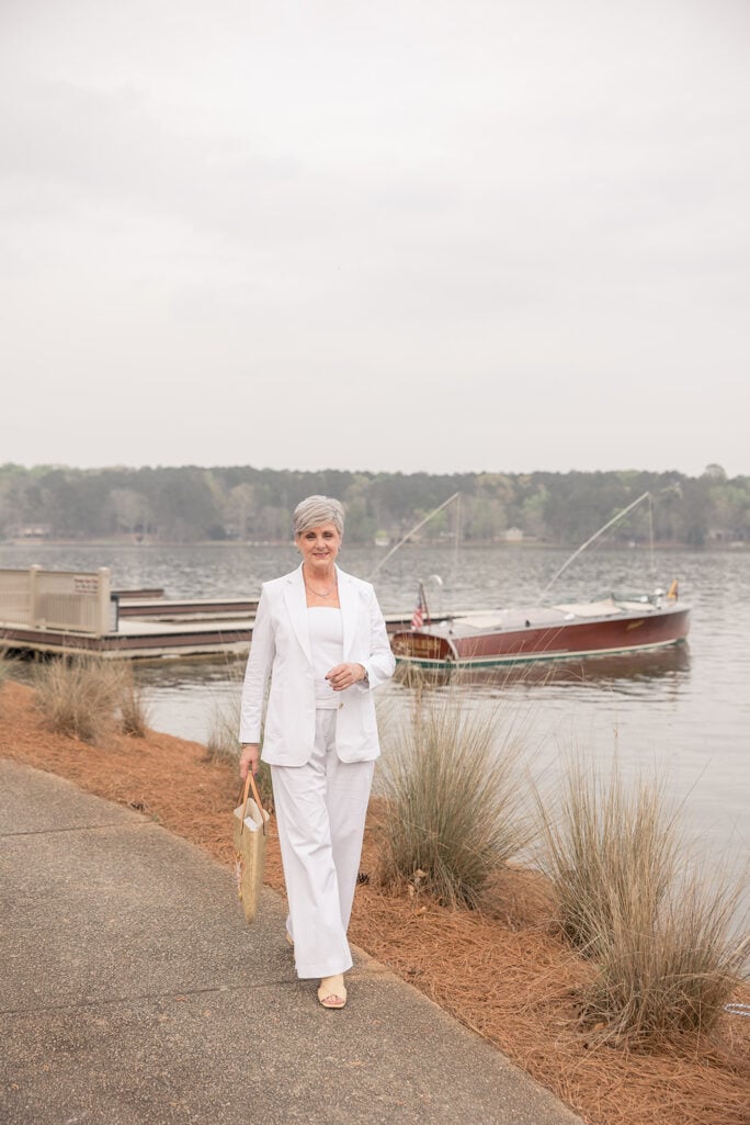 Woman over 50 with silver hair wearing a monochromatic all-white outfit featuring a white linen blazer, white tank top, and white wide-leg pants with nude heeled sandals, carrying a straw tote with floral embroidery at a lakeside dock — a summer white blazer outfit idea for women over 50.