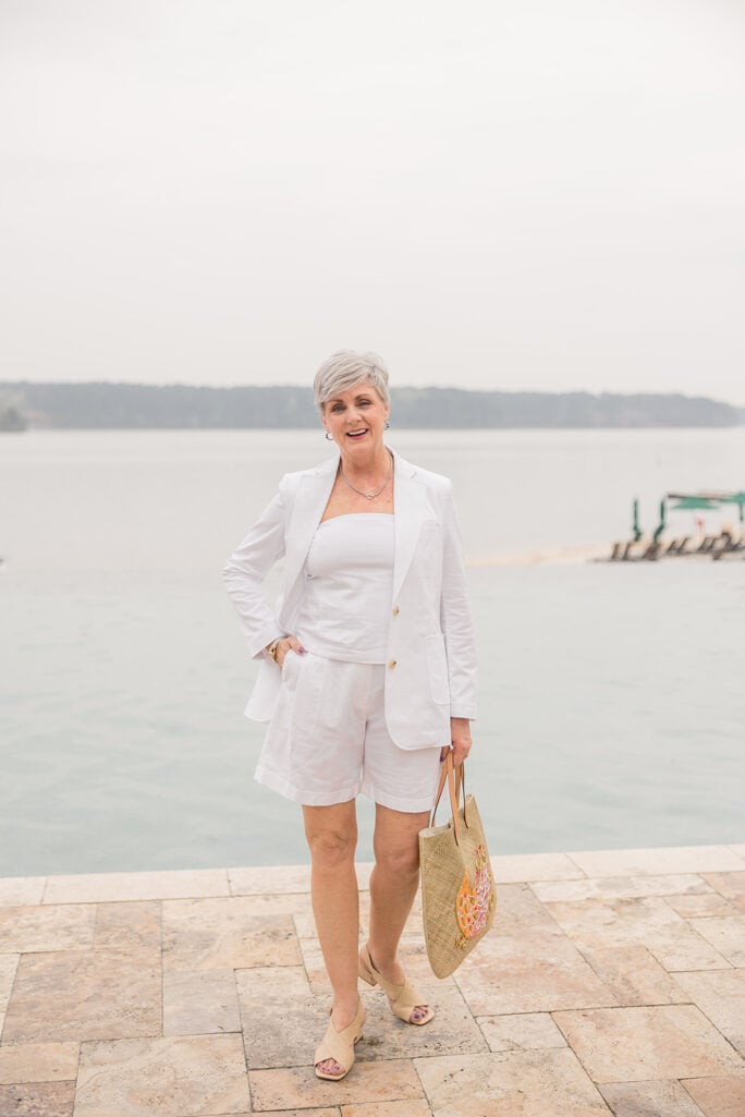 Woman over 50 with silver hair wearing a tailored white linen blazer over a white tank top with white tailored shorts and nude block heel sandals, carrying a straw tote with pink and orange floral embroidery on a stone patio by the water — a playful summer white blazer and shorts outfit for women over 50.