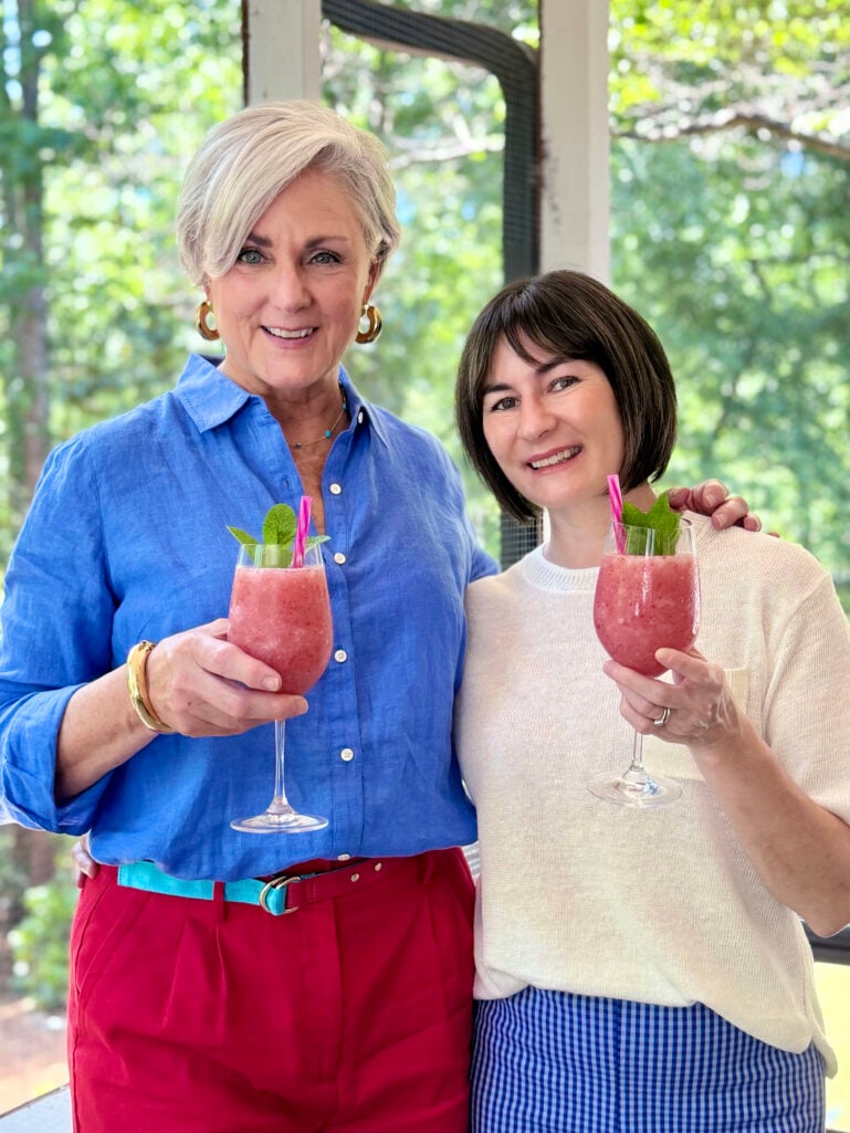Beth in a blue linen shirt and Kelly in a cream knit top smiling and holding up frozen strawberry frosé cocktails garnished with mint and pink straws on the screened porch.