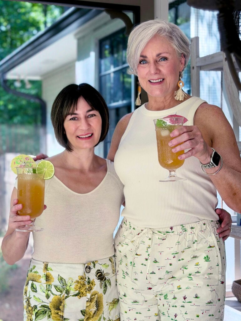 Kelly in a cream cashmere tee and Beth in a cream ribbed tank top with raffia tassel earrings, smiling and holding up tropical rum cocktails in stemmed hurricane glasses garnished with lime wheels, mint, and pink umbrellas on the screened porch.