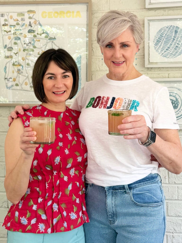 Kelly and Beth stand together smiling at the camera, each holding a rocks glass of spicy paloma with a tajín rim, Kelly in her red floral peplum top and Beth in her white Bonjour tee, the framed Georgia map print visible behind them.