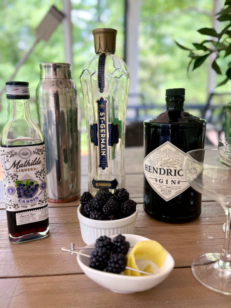 An outdoor table styled with a bottle of Mathilde Cassis liqueur, a bottle of St-Germain elderflower liqueur, a bottle of Hendrick's gin, a stainless cocktail shaker, a white bowl of fresh blackberries, and a small dish with blackberry and lemon twist garnishes on cocktail picks, green trees visible through the screened porch.