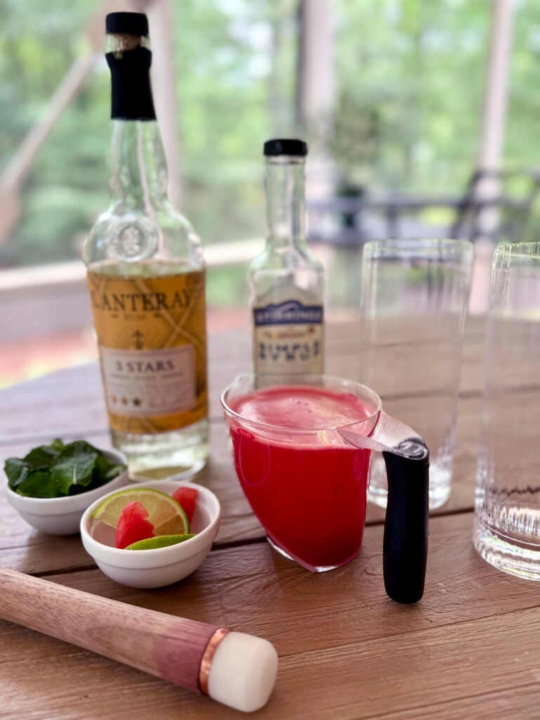 An outdoor table styled with a bottle of Plantation 3 Stars white rum, a bottle of simple syrup, a glass measuring cup filled with fresh blended watermelon juice, a small white bowl of fresh mint leaves, a white bowl of lime and watermelon garnishes, and a wooden muddler, green trees visible through the screened porch in the background.