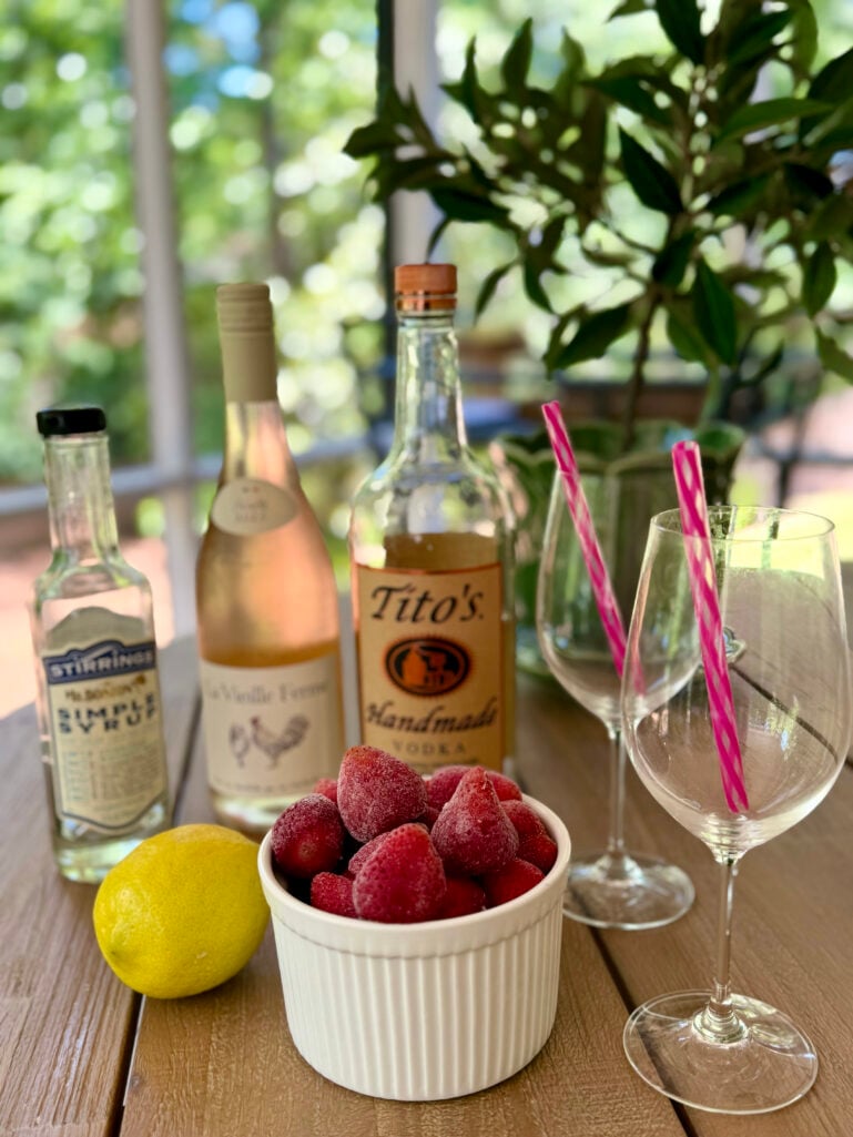 Frozen Frosé ingredients arranged on a wood table — a bottle of rosé, Tito's vodka, Stirrings simple syrup, a lemon, and a white ramekin of frozen strawberries, with two empty wine glasses and pink striped straws.