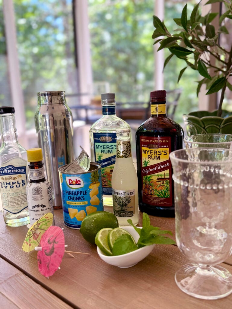 Tropical rum cocktail ingredients arranged on a wood table — Stirrings simple syrup, Angostura bitters, a silver cocktail shaker, a can of Dole pineapple chunks, Fever-Tree ginger beer, two bottles of Myers's rum (Platinum White and Original Dark), lime slices and fresh mint in a small white bowl, paper cocktail umbrellas, and an empty glass.