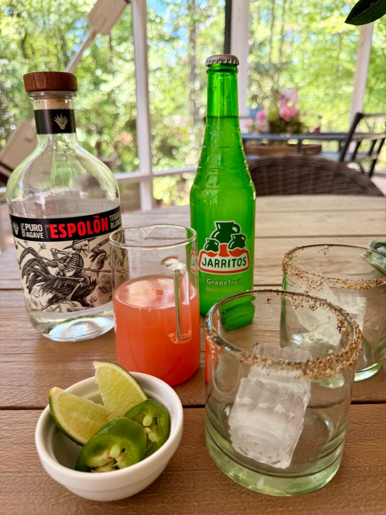 An outdoor table styled with a bottle of Espolón Blanco tequila, a bottle of Jarritos Grapefruit soda, a glass pitcher of pink grapefruit juice, two tajín-rimmed rocks glasses with large ice cubes, and a small white bowl of fresh jalapeño slices and lime wedges, lush green trees visible through the screened porch windows.