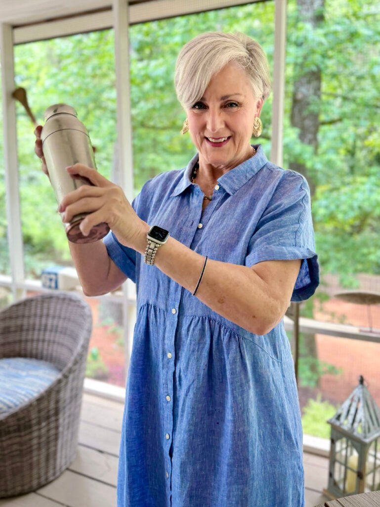 Beth stands on the screened porch smiling, shaking a stainless cocktail shaker with both hands to prepare the Blackberry French Gimlets, wearing her periwinkle blue linen shirtdress and gold jewelry, with wicker porch furniture and green trees visible behind her.