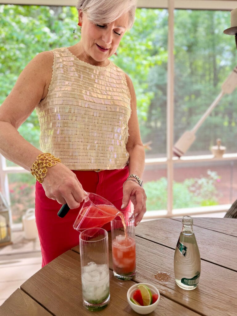 Beth leans over an outdoor table, pouring fresh blended watermelon juice from a glass measuring cup into two highball glasses filled with muddled mint and ice, wearing her cream sequin shell top and red wide-leg jeans, club soda and garnishes nearby on the table.
