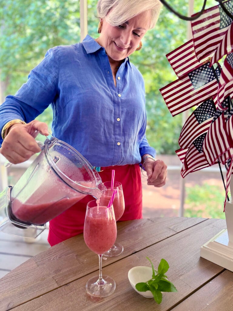 Beth in a blue linen shirt pouring blended strawberry frosé from a blender pitcher into stemmed wine glasses on the porch, with American flags and a sprig of fresh mint nearby.